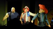 Three guys singing, one bald and big, one in star and strips, and one with big ears and hat
