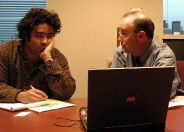 One guy talking to another in front of a laptop