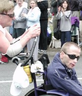 Father and son wearing sunglasses, with the older one leaning on the special handles of a sport wheel chair he is pushing