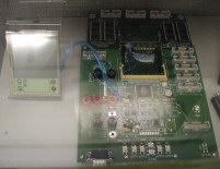 Large circuit board next to separate screen and buttons photographed inside a protective display case