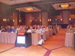 People sitting behind tables in a ballroom with a TV monitor with a big digital clock facing the stage