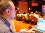 Dan photographed from the side as he types while looking at his laptop screen with rows of attendees in the distance