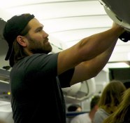 Serious Johnny arranging overhead luggage with black baseball hat on backwards