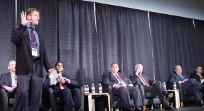 Mike standing, six people sitting in comfortable chairs on stage