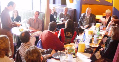 Long table and two booths of people looking at Richard standing and reading